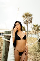 A woman in a black bikini poses near a rope fence with palm trees and foliage in the background. She has long dark hair and is wearing jewelry, including earrings, a bracelet, and a necklace. The atmosphere suggests a sunny, warm day, possibly at a beach or coastal area.