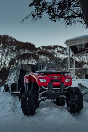 A bright red all-terrain vehicle (ATV) equipped with track wheels is parked in a snowy environment. Nearby, there is a metal cage and a trailer partially covered with snow. Sparse trees and an overcast sky form the background, along with a wooden structure with icicles hanging from the roof.
