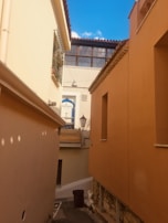 Narrow alleyway with tall, peach-colored and beige buildings on either side. A brightly lit blue sky with scattered white clouds above. A wooden door with the sign 'Al Hammam' written on it is visible ahead, along with a classic streetlamp. The alleyway is quiet with no visible people.