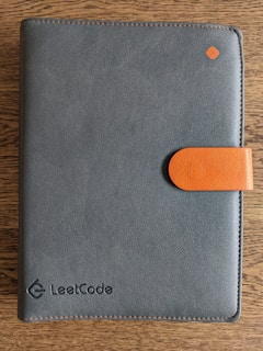 A dark gray leather notebook with an orange clasp lies on a wooden surface. The word 'LeetCode' is embossed on the lower right corner of the cover, and there is a small orange diamond-shaped detail near the top right corner.