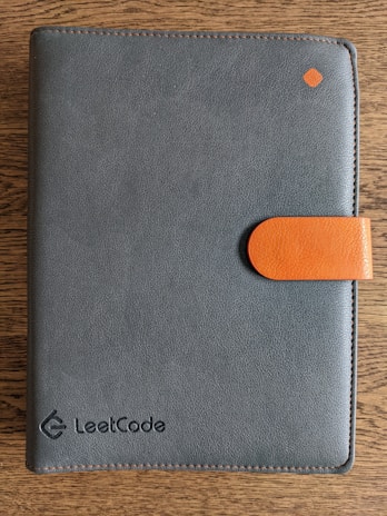 A dark gray leather notebook with an orange clasp lies on a wooden surface. The word 'LeetCode' is embossed on the lower right corner of the cover, and there is a small orange diamond-shaped detail near the top right corner.