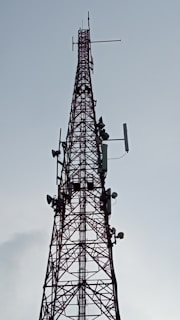 A tall, metal communication tower with multiple antennas and dishes attached, stretching vertically into the sky against a backdrop of a cloudy sky.
