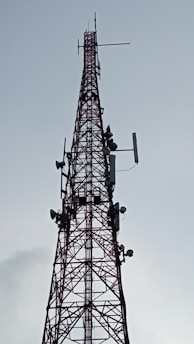 A tall, metal communication tower with multiple antennas and dishes attached, stretching vertically into the sky against a backdrop of a cloudy sky.