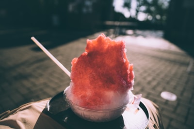 A vibrant red and white shaved ice dessert is served in a clear plastic bowl. It is sitting on a dark surface, likely a tray, with a wooden stick, possibly a spoon, inserted into it. The background is blurred, suggesting an outdoor setting with warm lighting and shadow patterns on a paved surface.