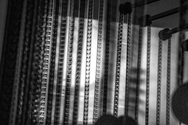 Black and white image displaying vertical lines with intricate patterns on a translucent surface. The lighting casts shadows and highlights, creating an abstract and artistic effect. Some cylindrical objects are present on the right side.