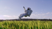 An astronaut in a white space suit is floating above a field of green grass. The sky is clear with a few scattered clouds, and there’s a distant line of trees in the background. The scene combines elements of space exploration with nature, creating a surreal and dreamlike atmosphere.