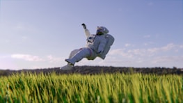 An astronaut in a white space suit is floating above a field of green grass. The sky is clear with a few scattered clouds, and there’s a distant line of trees in the background. The scene combines elements of space exploration with nature, creating a surreal and dreamlike atmosphere.