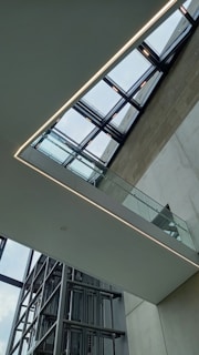 A modern architectural interior featuring a glass ceiling and a sleek staircase with glass railings. The space is characterized by clean lines and a minimalist design. The ceiling consists of a grid of windows allowing natural light to flood the space.