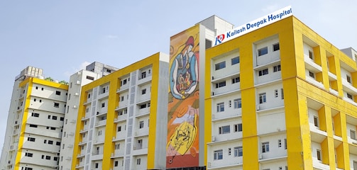 Several modern buildings with yellow and white facades stand under a clear sky. One of the buildings features a colorful mural, and another has a sign for Kailash Deepak Hospital.