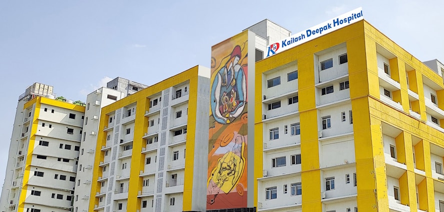 Several modern buildings with yellow and white facades stand under a clear sky. One of the buildings features a colorful mural, and another has a sign for Kailash Deepak Hospital.