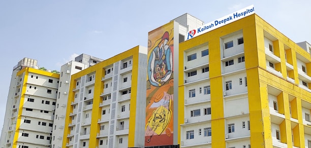 Several modern buildings with yellow and white facades stand under a clear sky. One of the buildings features a colorful mural, and another has a sign for Kailash Deepak Hospital.