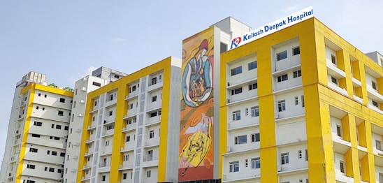 Several modern buildings with yellow and white facades stand under a clear sky. One of the buildings features a colorful mural, and another has a sign for Kailash Deepak Hospital.
