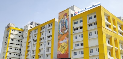 Several modern buildings with yellow and white facades stand under a clear sky. One of the buildings features a colorful mural, and another has a sign for Kailash Deepak Hospital.