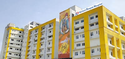 Several modern buildings with yellow and white facades stand under a clear sky. One of the buildings features a colorful mural, and another has a sign for Kailash Deepak Hospital.