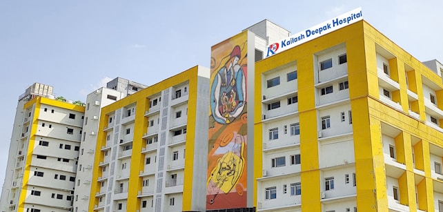 Several modern buildings with yellow and white facades stand under a clear sky. One of the buildings features a colorful mural, and another has a sign for Kailash Deepak Hospital.