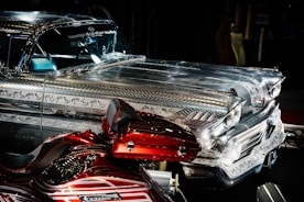 A highly detailed vintage car with intricate metal engravings and patterns on the bodywork. The vehicle has a shiny, reflective surface that contrasts with a deep red, custom-painted motorcycle featuring elaborate designs. Both vehicles are displayed prominently in a dimly lit environment, emphasizing their craftsmanship.