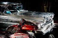 A highly detailed vintage car with intricate metal engravings and patterns on the bodywork. The vehicle has a shiny, reflective surface that contrasts with a deep red, custom-painted motorcycle featuring elaborate designs. Both vehicles are displayed prominently in a dimly lit environment, emphasizing their craftsmanship.