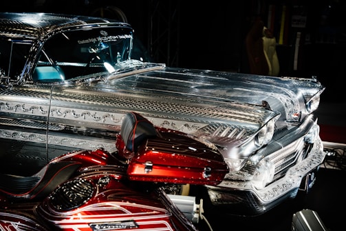 A highly detailed vintage car with intricate metal engravings and patterns on the bodywork. The vehicle has a shiny, reflective surface that contrasts with a deep red, custom-painted motorcycle featuring elaborate designs. Both vehicles are displayed prominently in a dimly lit environment, emphasizing their craftsmanship.