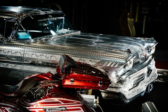 A highly detailed vintage car with intricate metal engravings and patterns on the bodywork. The vehicle has a shiny, reflective surface that contrasts with a deep red, custom-painted motorcycle featuring elaborate designs. Both vehicles are displayed prominently in a dimly lit environment, emphasizing their craftsmanship.