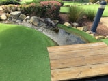 A small artificial stream bordered by rocks flows through a landscape of manicured green turf. Bushes with pink flowers and tall grasses grow nearby, adding to the natural setting. A wooden deck or bridge is in the foreground, and a tall lamp post stands on the right side.