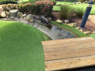 A small artificial stream bordered by rocks flows through a landscape of manicured green turf. Bushes with pink flowers and tall grasses grow nearby, adding to the natural setting. A wooden deck or bridge is in the foreground, and a tall lamp post stands on the right side.