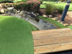 A small artificial stream bordered by rocks flows through a landscape of manicured green turf. Bushes with pink flowers and tall grasses grow nearby, adding to the natural setting. A wooden deck or bridge is in the foreground, and a tall lamp post stands on the right side.