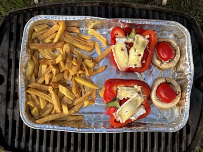 A foil tray containing cooked fries, stuffed bell peppers, and stuffed mushrooms is placed on a grill. The fries are golden brown, while the bell peppers are red and topped with slices of cheese. The mushrooms have bright red centers, likely tomatoes.
