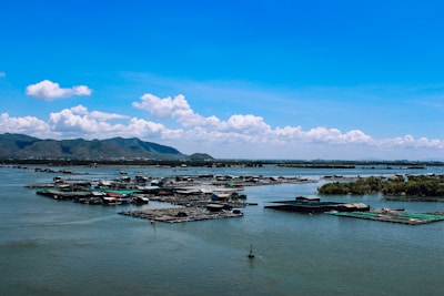 A serene waterbody with numerous floating structures, possibly fish farms or floating houses, spreads across the scene. The distant horizon is lined with lush green hills under a bright blue sky dotted with fluffy white clouds.