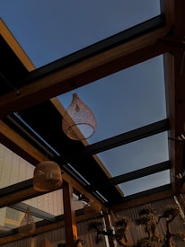 A rooftop view featuring wooden beams and glass panels in a modern architectural style. Woven rattan pendant lights hang from the ceiling, creating a cozy and rustic atmosphere. The sky is visible through the glass, indicating a clear day.