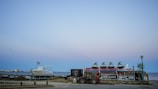 At the seaside, multiple jet skis are stacked on a trailer next to a parked boat with a distant shoreline and buildings visible under a clear sky. There are signs for rental services on the trailer, and a lamp post nearby.