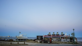 At the seaside, multiple jet skis are stacked on a trailer next to a parked boat with a distant shoreline and buildings visible under a clear sky. There are signs for rental services on the trailer, and a lamp post nearby.