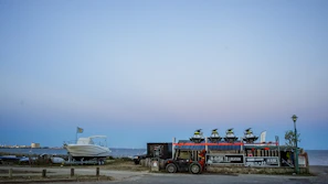 At the seaside, multiple jet skis are stacked on a trailer next to a parked boat with a distant shoreline and buildings visible under a clear sky. There are signs for rental services on the trailer, and a lamp post nearby.