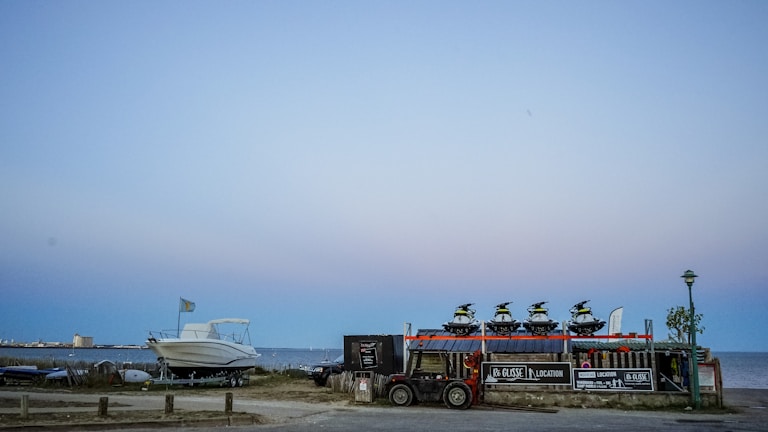 At the seaside, multiple jet skis are stacked on a trailer next to a parked boat with a distant shoreline and buildings visible under a clear sky. There are signs for rental services on the trailer, and a lamp post nearby.