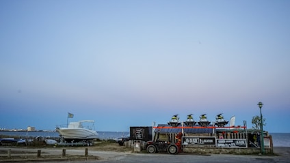 At the seaside, multiple jet skis are stacked on a trailer next to a parked boat with a distant shoreline and buildings visible under a clear sky. There are signs for rental services on the trailer, and a lamp post nearby.