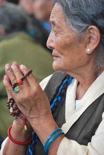 An elderly woman with gray hair braided with a blue thread, wearing traditional clothing and holding prayer beads. She has a contemplative expression and wears colorful bracelets and a ring with a turquoise stone.