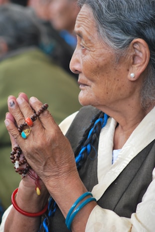 An elderly woman with gray hair braided with a blue thread, wearing traditional clothing and holding prayer beads. She has a contemplative expression and wears colorful bracelets and a ring with a turquoise stone.