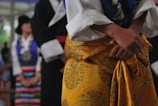 A person wearing traditional clothing dominates the foreground. The attire includes a yellow garment with intricate patterns, complemented by a white shirt with wide sleeves. In the background, other individuals are visible, also dressed in colorful traditional outfits, one featuring stripes.