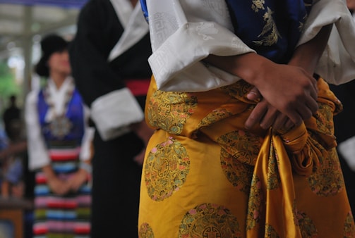 A person wearing traditional clothing dominates the foreground. The attire includes a yellow garment with intricate patterns, complemented by a white shirt with wide sleeves. In the background, other individuals are visible, also dressed in colorful traditional outfits, one featuring stripes.
