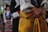 A person wearing traditional clothing dominates the foreground. The attire includes a yellow garment with intricate patterns, complemented by a white shirt with wide sleeves. In the background, other individuals are visible, also dressed in colorful traditional outfits, one featuring stripes.