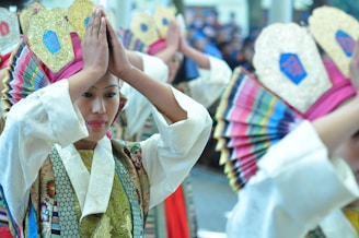 A group of people dressed in traditional, colorful attire with intricate patterns and embellishments are performing a dance. They are holding decorative fans and wearing headpieces adorned with gold and bright fabric.
