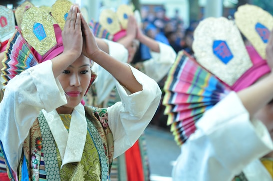 A group of people dressed in traditional, colorful attire with intricate patterns and embellishments are performing a dance. They are holding decorative fans and wearing headpieces adorned with gold and bright fabric.