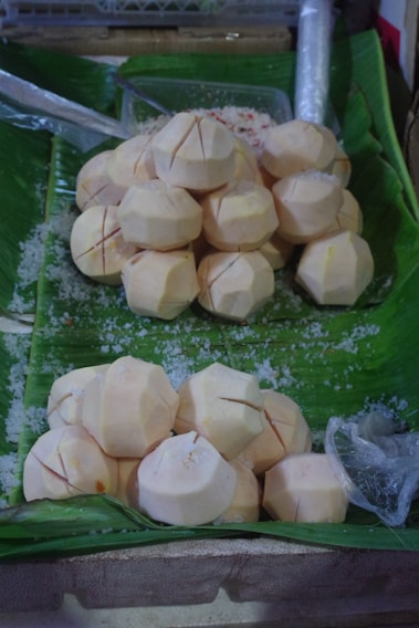 Spherical fruits or vegetables with a peeled and clean surface are arranged on large green banana leaves. The items have been scored with small cuts, and salt is scattered around them, suggesting a preparation or preservation process. In the background, plastic wrap and a metal spoon are visible.