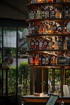 A well-organized display of various bottles of alcohol stacked on a circular shelving unit in a bar setting. The background features large glass windows with lush greenery visible, and soft lighting creates an inviting and warm ambiance.