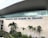 The modern facade of an airport features large glass windows and a wide stone wall with the name 'Aeropuerto José Joaquín de Olmedo' prominently displayed. In front of the building, several tropical plants and palm trees are arranged alongside a decorative water feature.
