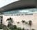 The modern facade of an airport features large glass windows and a wide stone wall with the name 'Aeropuerto José Joaquín de Olmedo' prominently displayed. In front of the building, several tropical plants and palm trees are arranged alongside a decorative water feature.