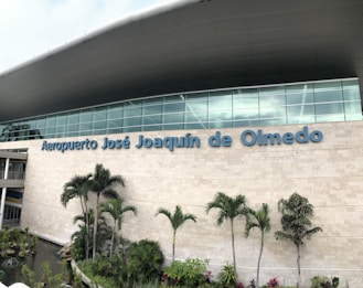The modern facade of an airport features large glass windows and a wide stone wall with the name 'Aeropuerto José Joaquín de Olmedo' prominently displayed. In front of the building, several tropical plants and palm trees are arranged alongside a decorative water feature.