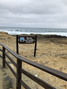 A wooden sign with the words 'Mirador La Puntilla Viewpoint' stands near a rugged coastline. Behind the sign, there is a view of the ocean with waves crashing against the shore under a cloudy sky. The area around the sign is sandy and rocky, and there is a wooden railing leading to the viewpoint.