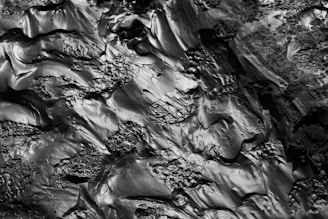 A textured surface with smooth and rough sections, resembling solidified lava or mud. The surface has a shiny, reflective quality with various organic shapes and creases.