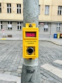 A yellow pedestrian crossing button mounted on a metal pole in an urban setting. The text on the button is in Czech, reading 'Čekejte' and 'Tlačítko pro chodce'. The background shows a cobblestone street and a building with windows.