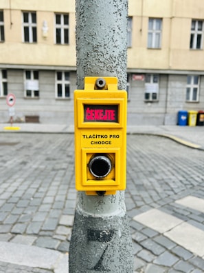 A yellow pedestrian crossing button mounted on a metal pole in an urban setting. The text on the button is in Czech, reading 'Čekejte' and 'Tlačítko pro chodce'. The background shows a cobblestone street and a building with windows.